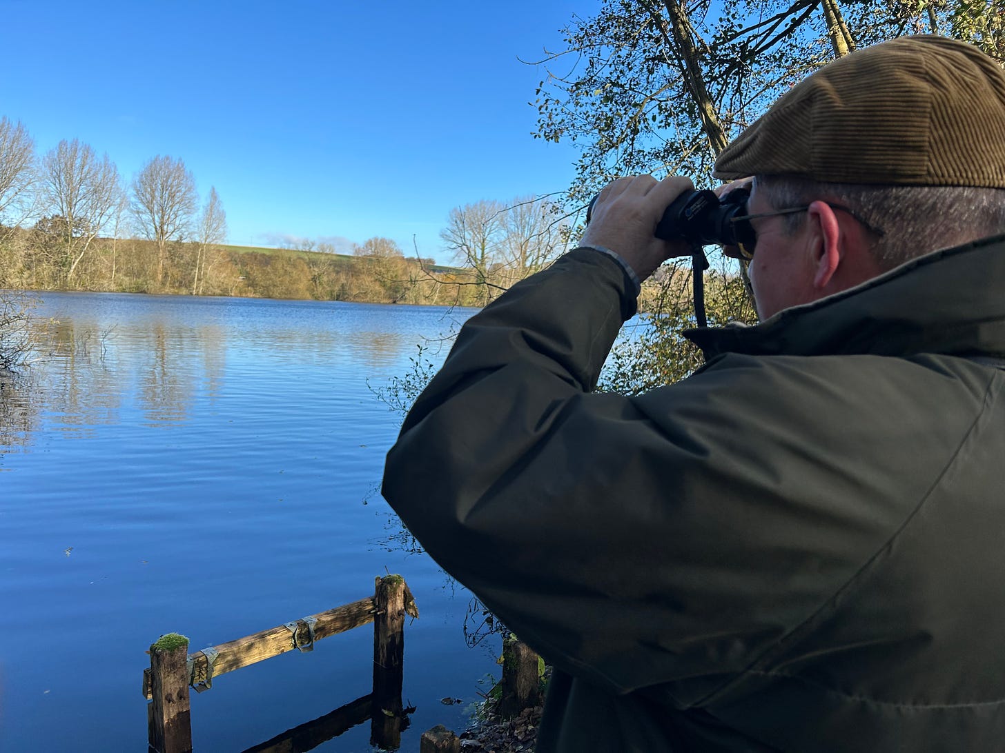 A man with binoculars looking over a lake at Langford Lakes, Steeple Langford, Wiltshire. The lakes are managed by the Wiltshire Wildlife Trust. Image: Roland's Travels