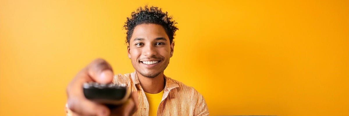 Smiling man holding a TV remote, against a bright yellow background. Caption: Bright smiles and ready to watch the Christmas Eve video by Blackmore's Night.