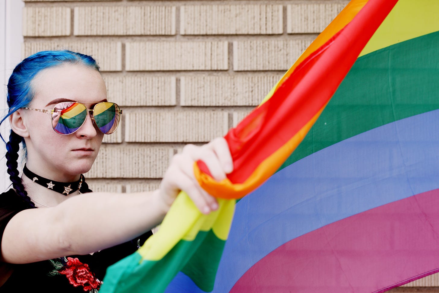 A young white woman with blue hair and mirror shades holding a pride flag. She also has a dog collar on.