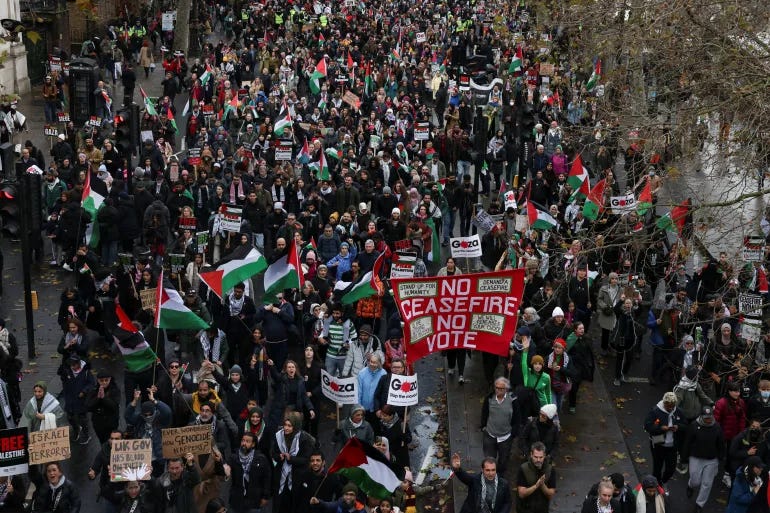 Color photo of protesters in London. In the center people hold a red banner that says "NO CEASEFIRE NO VOTE". There are also many Palestinian flags being waved and many homemade and printed signs. People look as if they are dressed for cold weather and the streets look slightly wet as if it had rained recently.