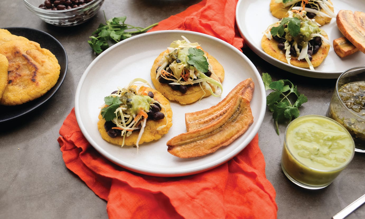 Vegetarian pupusas with black beans, curtido slaw and green salsa on top, on a plate with a side of plantains, on top of a red cloth napkin on a gray tabletop