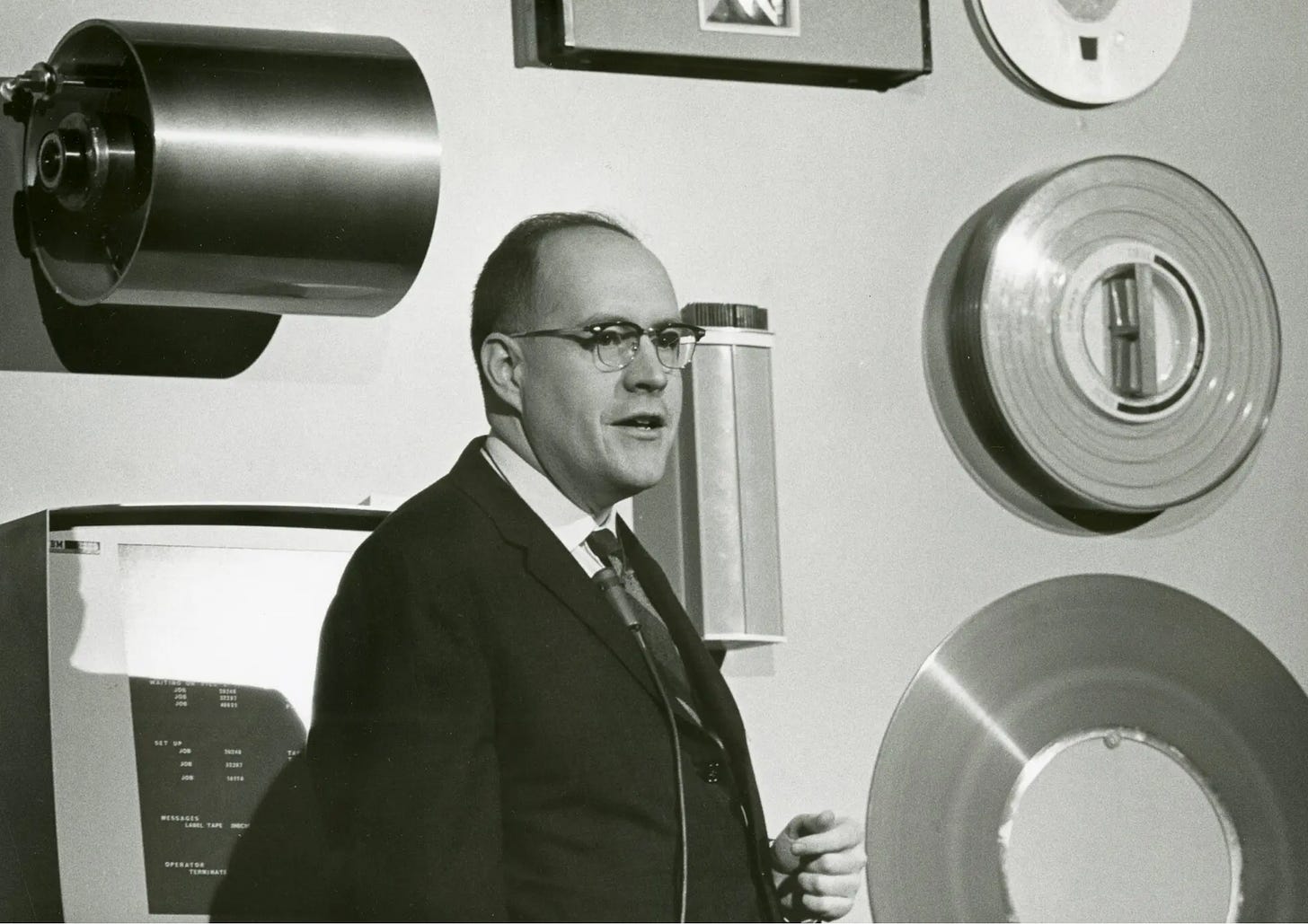 Black and white photo from the 1960s of a man in a suit delivering a speech with a background of then vintage computer components. 