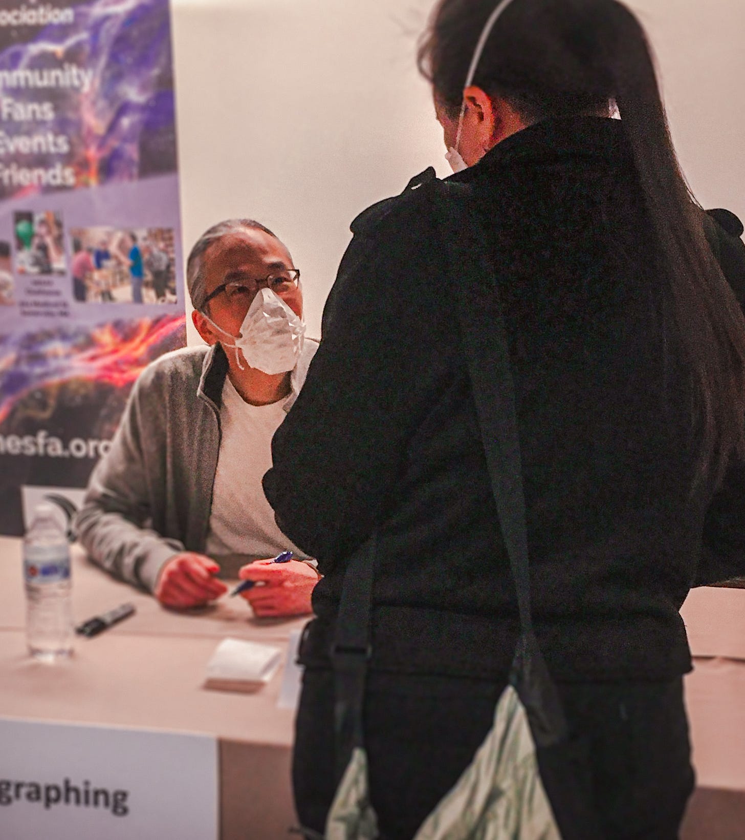 Um homem com ascendência chinesa, cabelos brancos e vestindo óculos e máscara, escuta uma mulher branca com cabelos lisos de costas. No cenário, uma mesa de autógrafo e um banner.