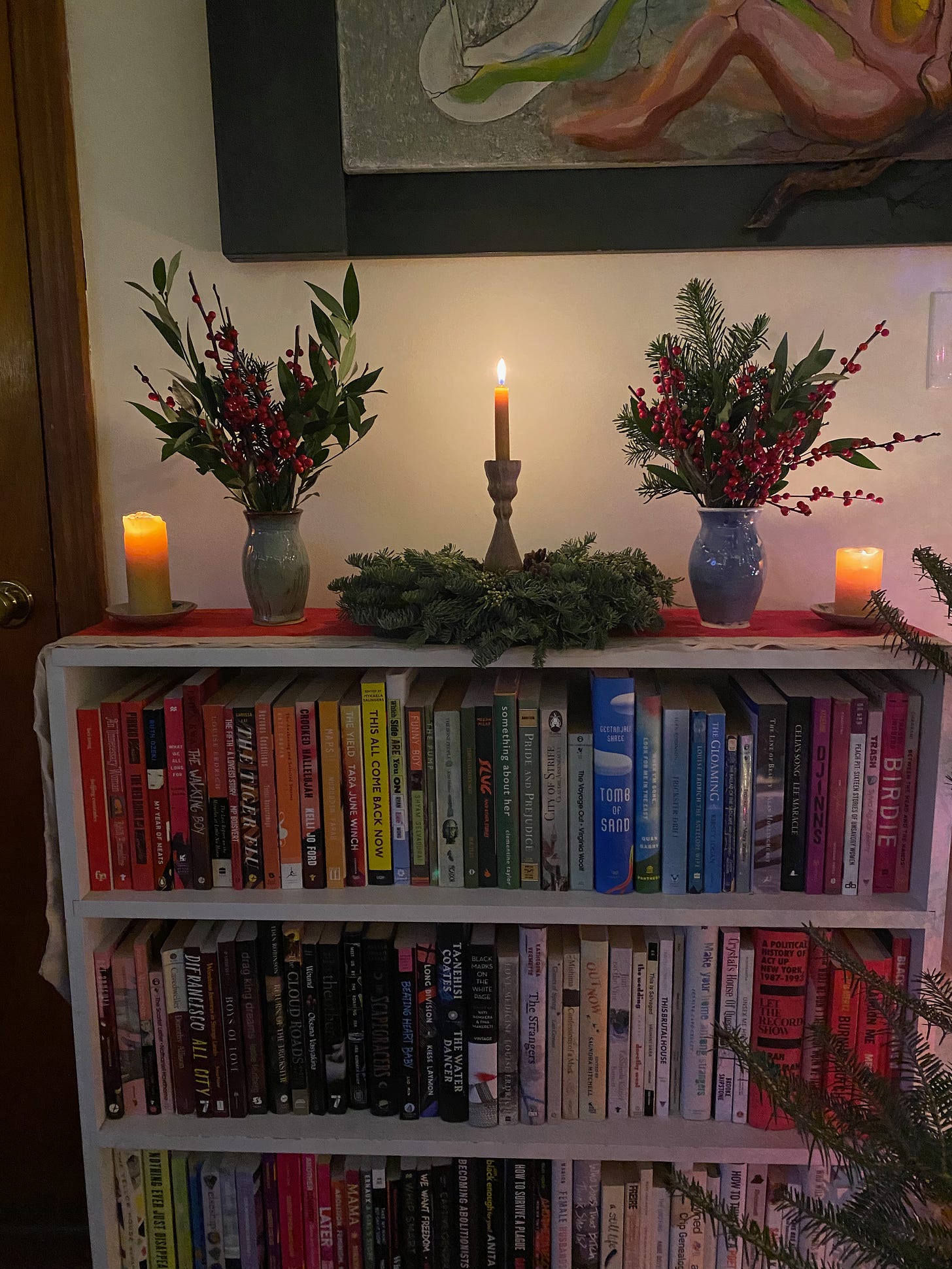 Several lit candles and jars of winterberry and greenery on top of a bookshelf.