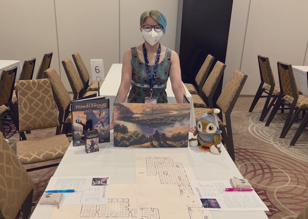 An image of Sebastian standing behind a Humblewood GM screen. There are character sheets and dice visible, as well as a Humblewood game book and owl plush toy.