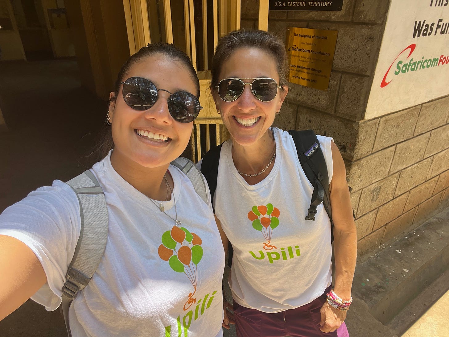 Carla and her daughter wear white tshirts that say 'upili'. Both wear singlasses, have brown hair and smile at the camera