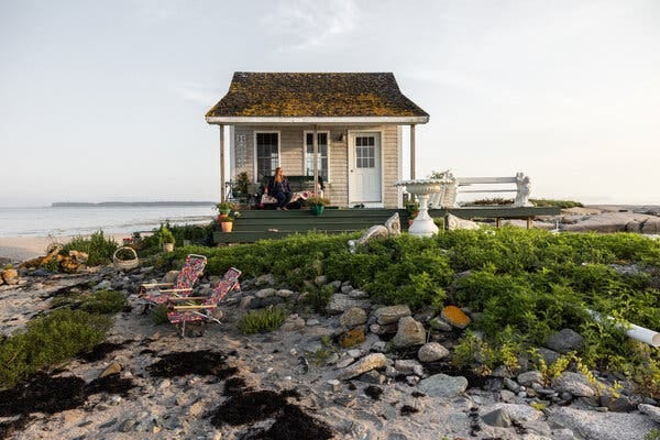 A tiny cabin sits on a rocky island off the coast of Maine. The island has a scrubby growth of green vegetation.