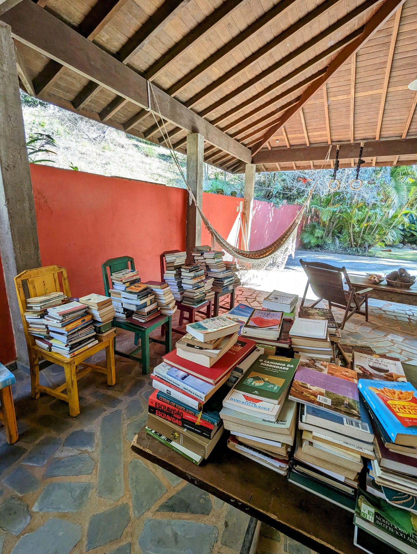 A covered area, with tables, chairs and a hammock, and plenty of greenery and sunshine beyond it. On the table and chairs are piled various books, ready to be wiped clean before being returned to the guesthouse's library.