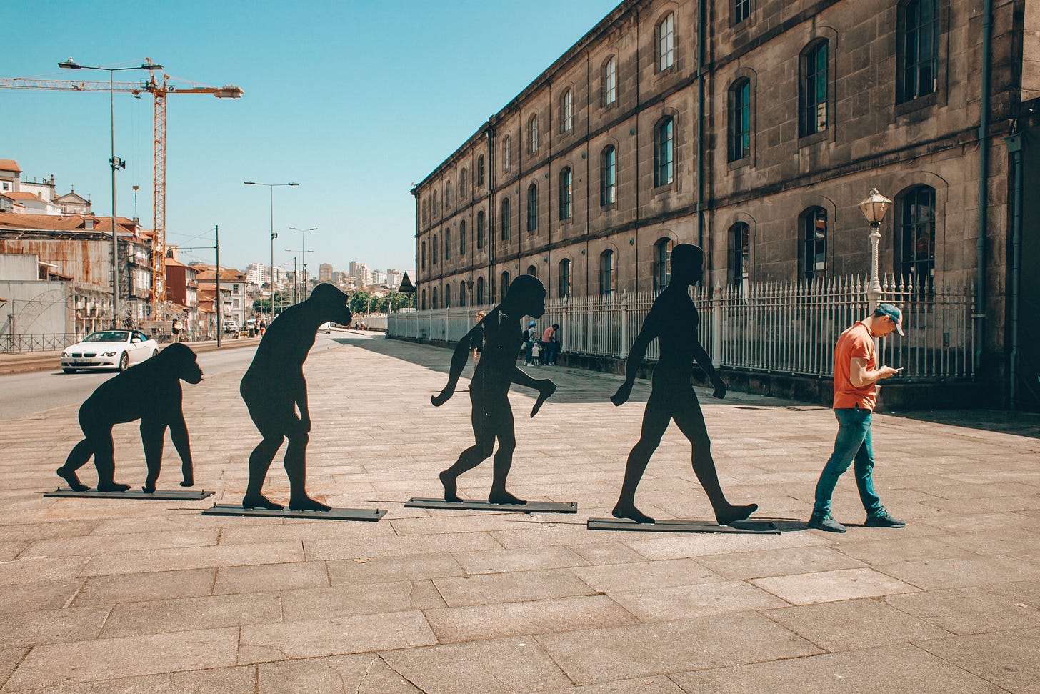 Statues on a sidewalk showing the evolution from ape to human while the last is a real human looking down on his mobile phone