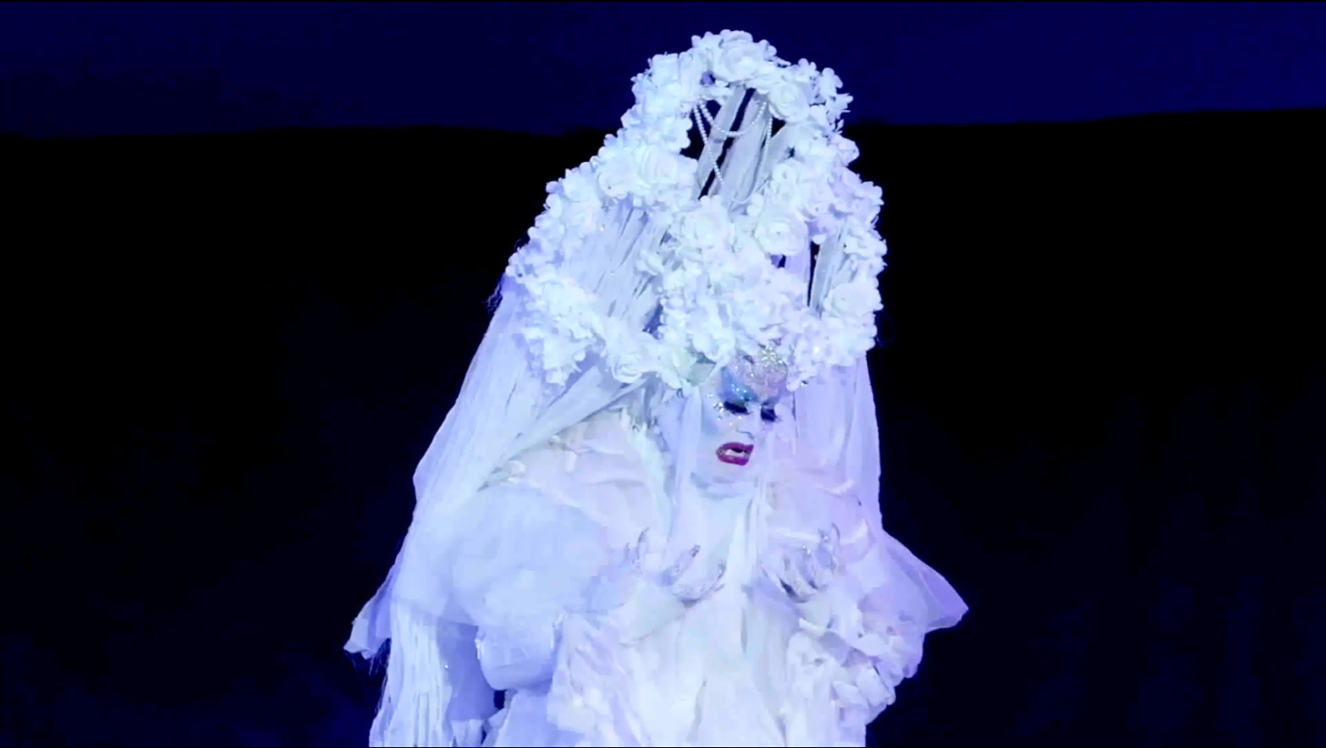 On a dark stage, a drag performer in an all-white ethereal, flowing ghost-like gown with an elaborate white-flower-and-ribbon crown. Their face and hands are also all white, with rhinestones adoring the face and fingers, with streaks of icey blue eyeshadow and magenta-red lips. They are looking down at their hands with an anguished expression.