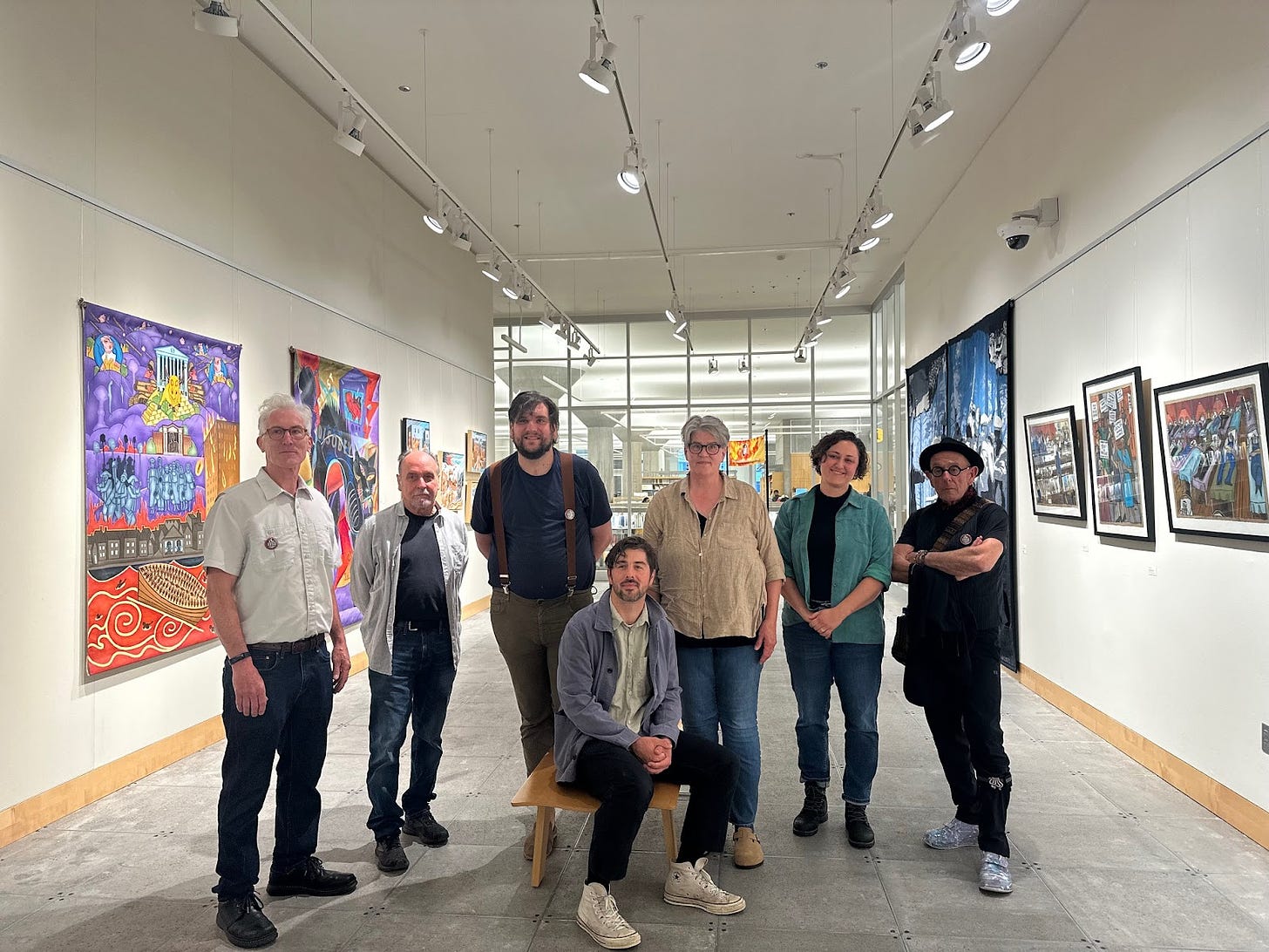 7 people pose in the middle of an art gallery with white walls and lights shining down, one man with a blue jacket and white sneakers sits in the middle on a bench