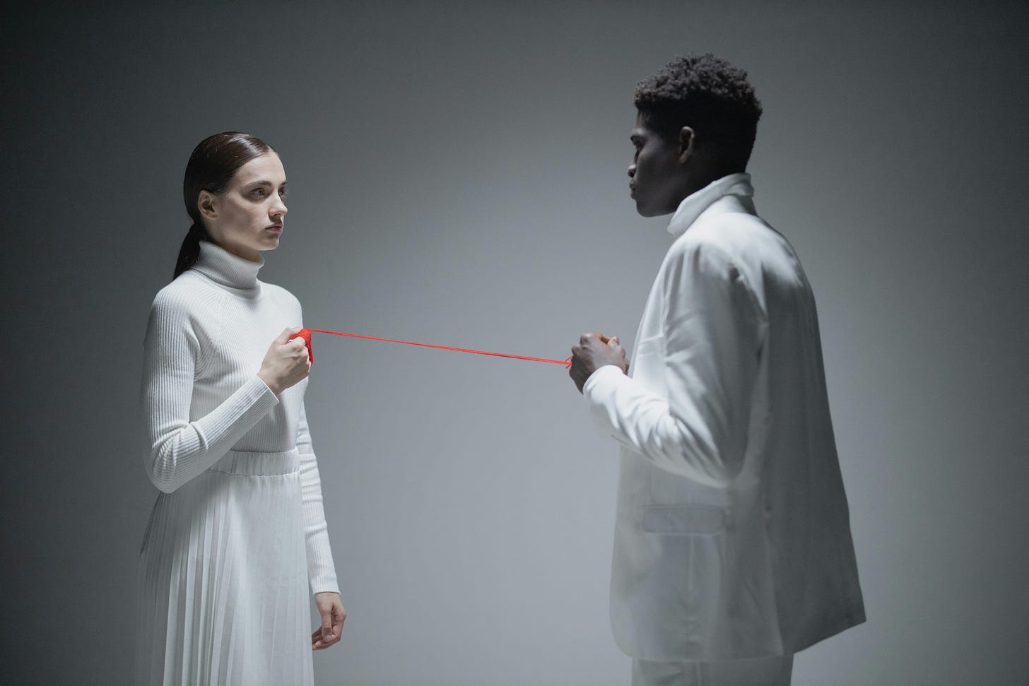Photo of white woman and black man wearing white clothes facing each other without smiling while holding a red threat between them at a distance of about 4 feet.