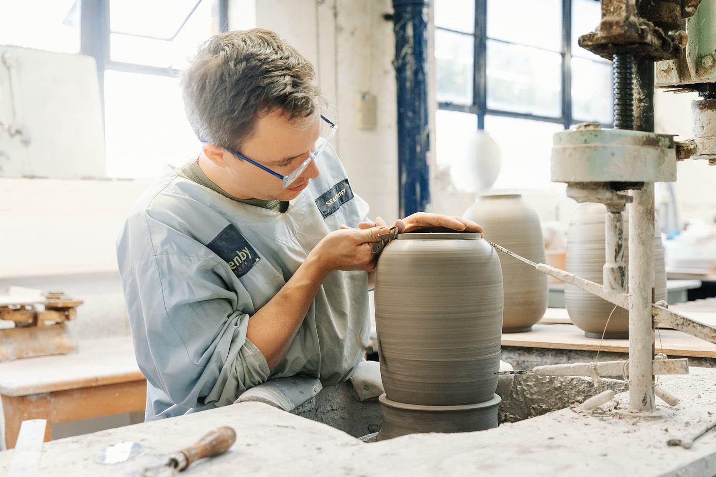 a young man is trimming a large clay vase in a factory or workshop setting. there is a huge window behind him and he is surrounded by other vases and machinery