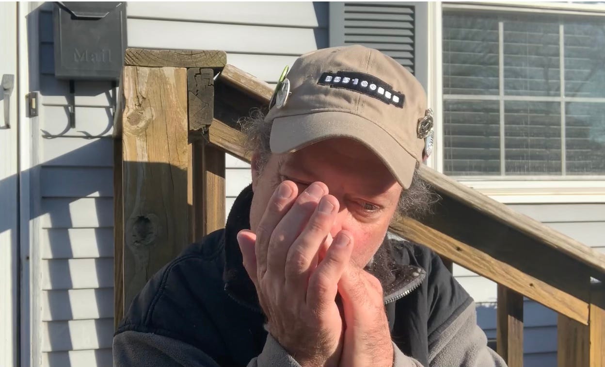 Gerard Mclean playing the harmonica on a front porch, wearing a tan hat with the 100 Harmonicas logo embroidered in the front, various buttons pinned to the sides. It is a bright, sunny morning