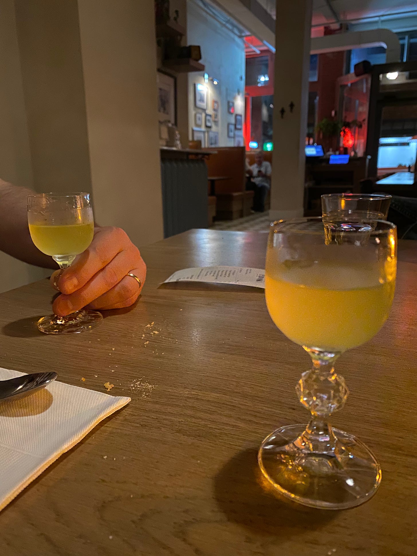 Two small glasses of limoncello on a table that's got crumbs and a napkin on it. Jeff's hand is holding the glass on the left side of the frame.