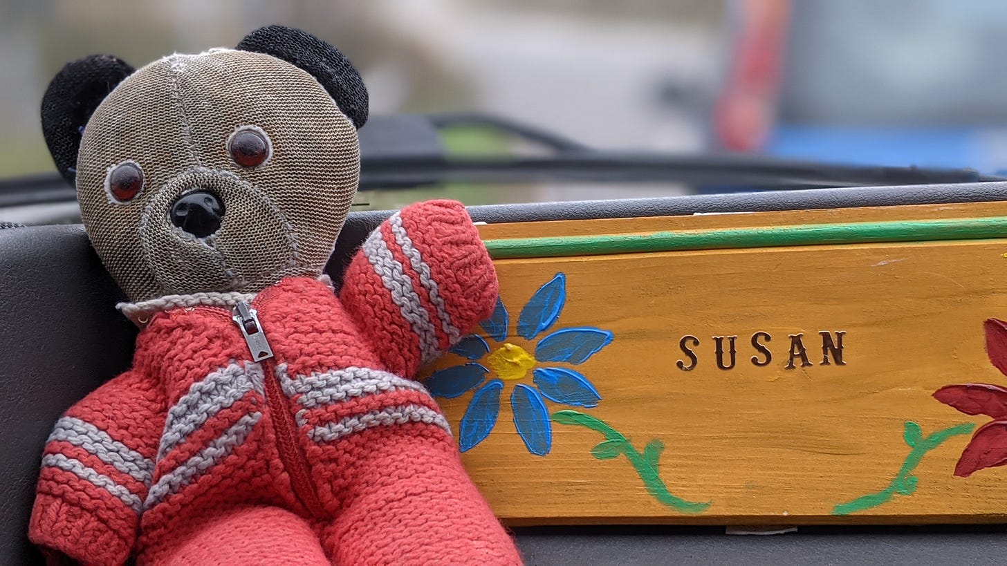 A well-loved old panda bear toy - dressed in a red jumpsuit - poses for the camera next to a wooden sign with hand-painted flowers and the name 'Susan' written in stickers.