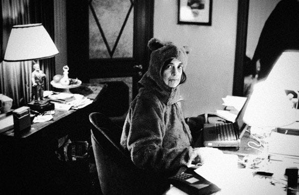 foto em preto e branco da autora Susan Sontag (mulher branca de cabelo e olhos castanhos) vestida com fantasia de urso em frente a uma mesa de trabalho