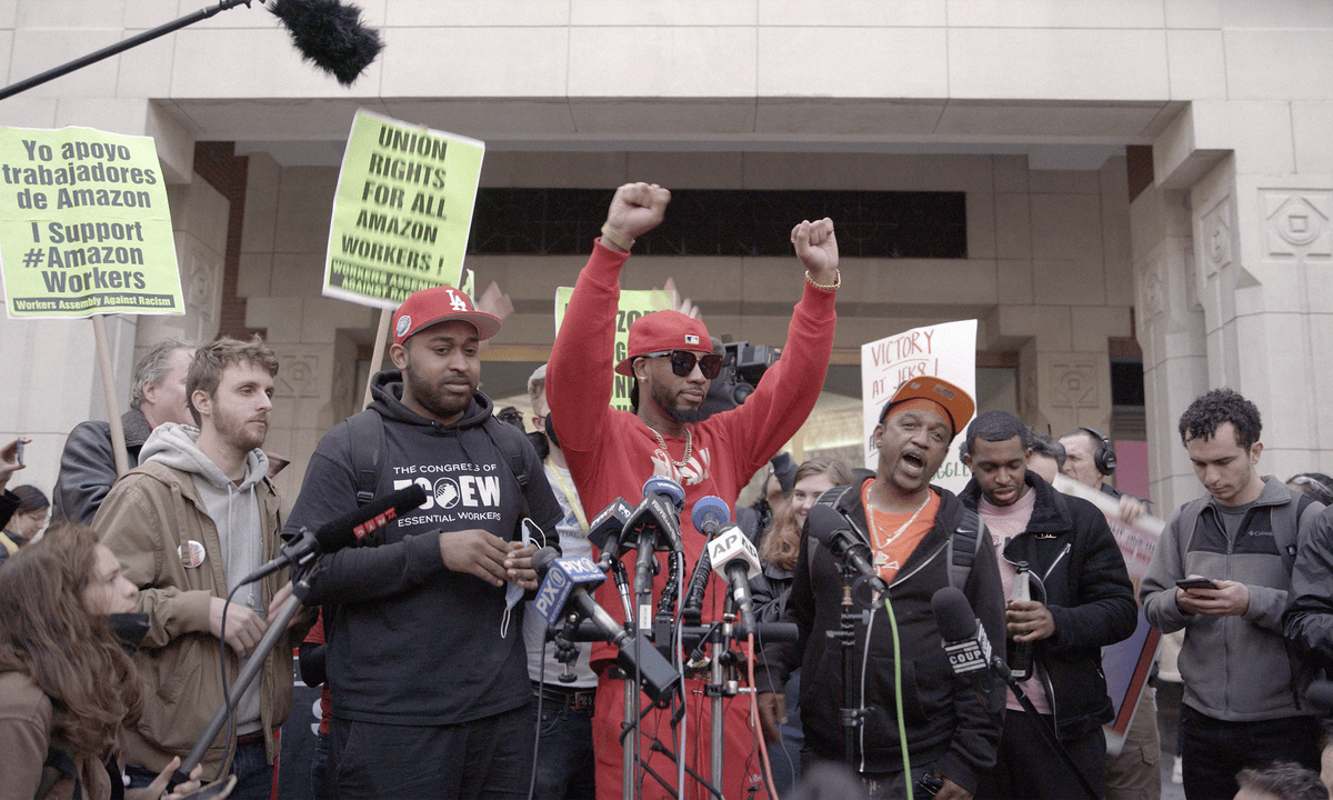 A shot of a press conference from the Amazon strike with Christian Smalls holding his arms up in victory.