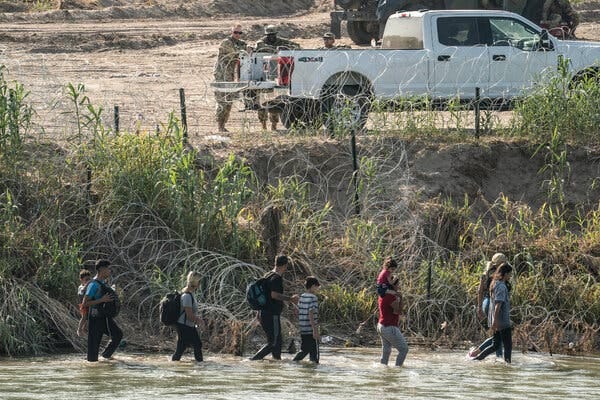 People wade along the edge of a river near a concertina wire barrier as soldiers in camouflage uniform watch from a pickup truck on the other side of the border.