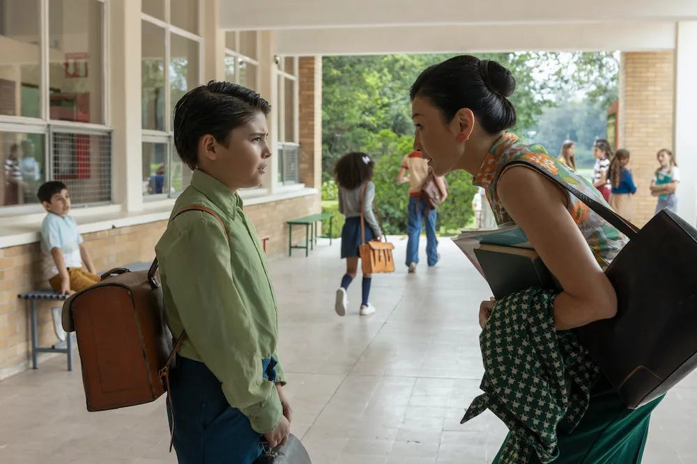 Foto de um garoto com uma mochila de couro antiga nas costas e sua professora inclinada para falar à altura dos olhos dele. Ela tem uma bolsa e casaco pendurados e muitos livros na mão. 