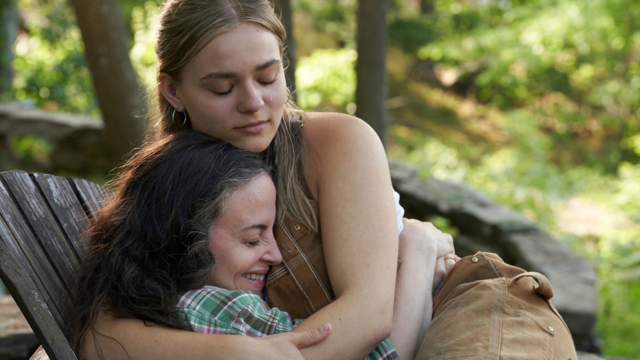 Two women sit in a deck chair in an outdoor setting. They smile and cry while hugging. 