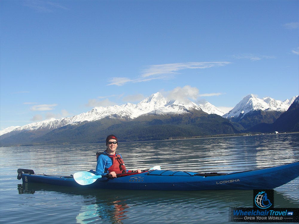 John on a kayak in the middle of a bay with snow-capped mountains in the background.
