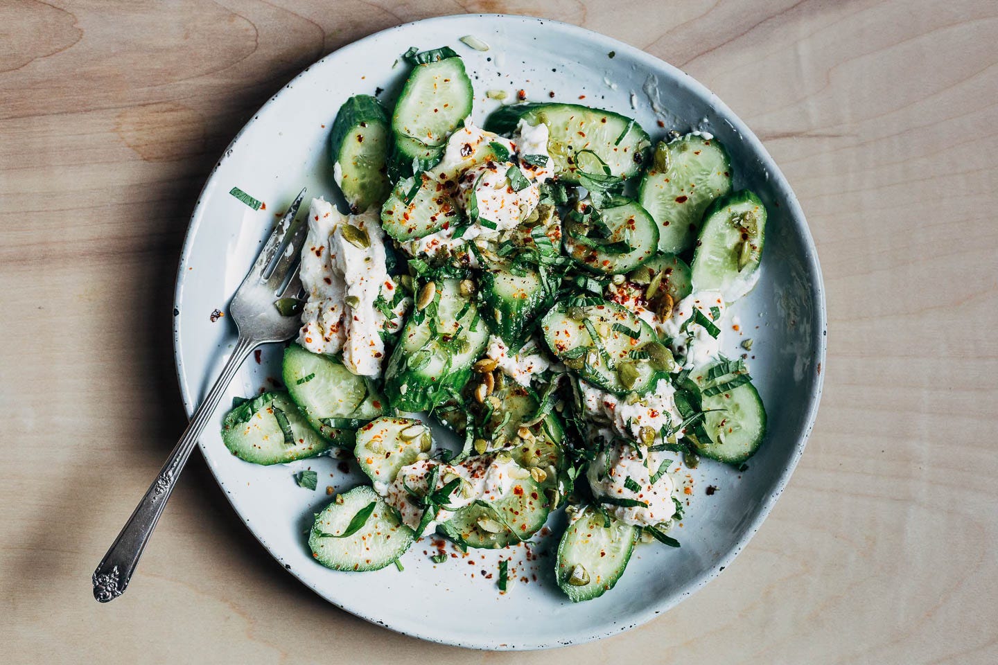 A salad with cucumbers and ricotta on a plate with herbs and pepper flakes. 