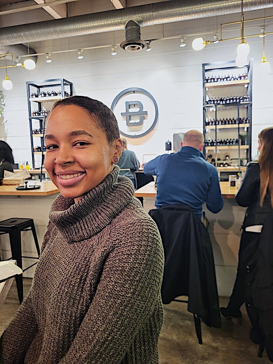 A woman smiling at the camera in a sage green cowl neck sweater and behind her are shelves of scented oils, a B logo, and people sitting at a bar mixing oil and wax to make candles with their backs to the camera