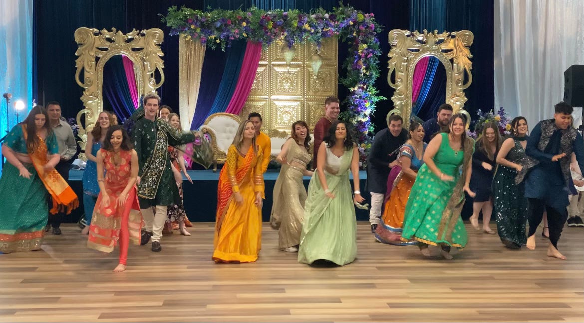 A group of men and women, in their 20s and 30s, dances wearing traditional Indian attire