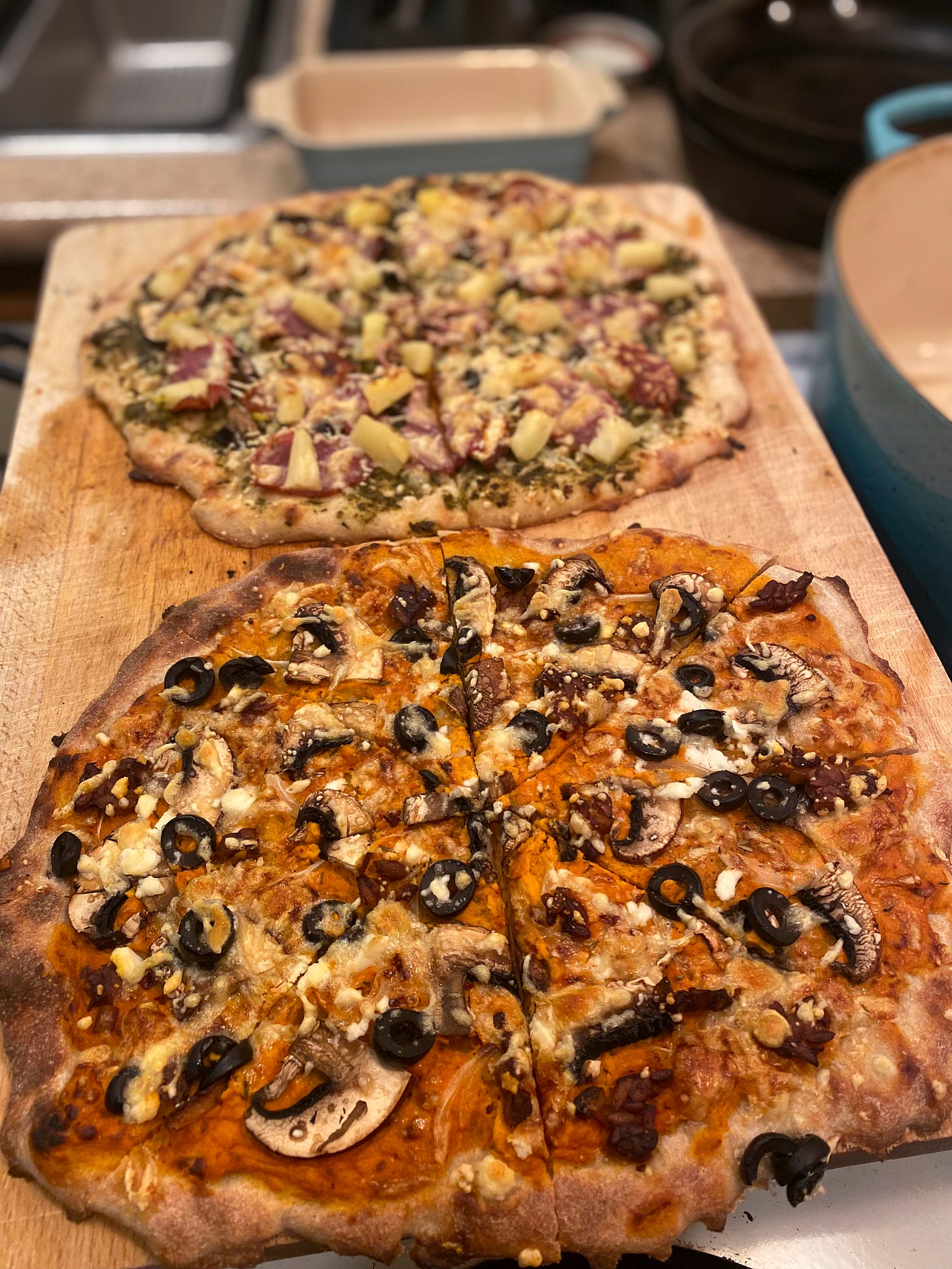 Two pizzas on a wooden cutting board. The nearest has an orange base sauce with olives and mushrooms, and the one in the background has pesto, pineapple, and capocollo.