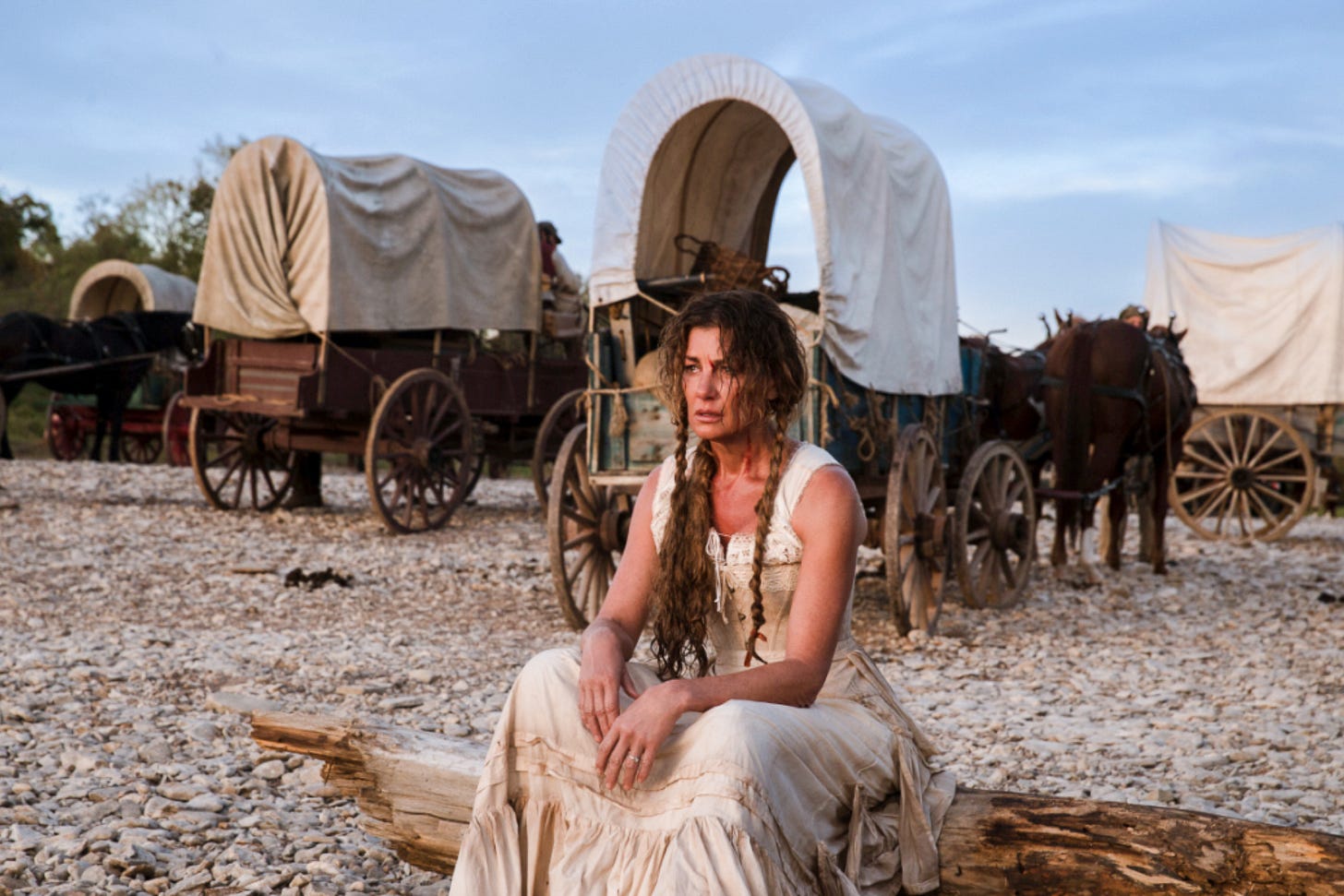 The scene from the series “1883” shows several horse-drawn covered wagons (known as prairie schooners) on a rocky riverbank. In the foreground, Margaret Dutton (played by Faith Hill) sits exhausted and pensive on a tree trunk. Her hair is disheveled, her dress is dirty – she embodies the harsh everyday life of the pioneer era. The scene from the series “1883” shows several horse-drawn covered wagons (known as prairie schooners) on a rocky riverbank. In the foreground, Margaret Dutton (played by Faith Hill) sits exhausted and pensive on a tree trunk. Her hair is disheveled, her dress is dirty – she embodies the harsh everyday life of the pioneer era.