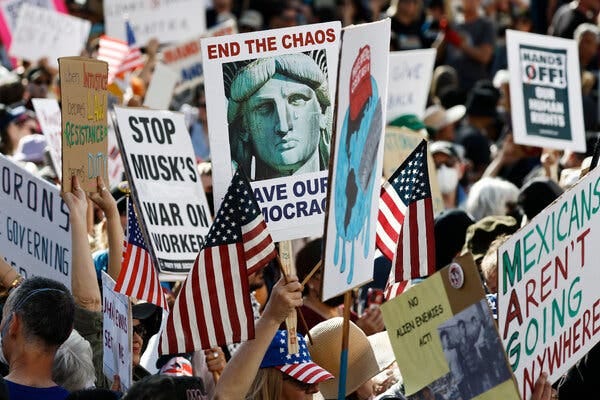 A crowd of protesters carrying American flags and signs denouncing Trump administration policies.