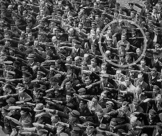 one man in a crowd making the nazi salute stands with his arms folded, defiant