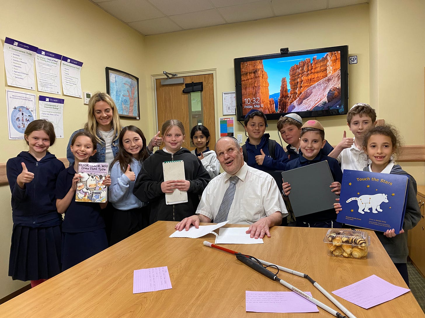 Rabbi Michael Levy sits at a table in a classroom surrounded by smiling students and their teacher