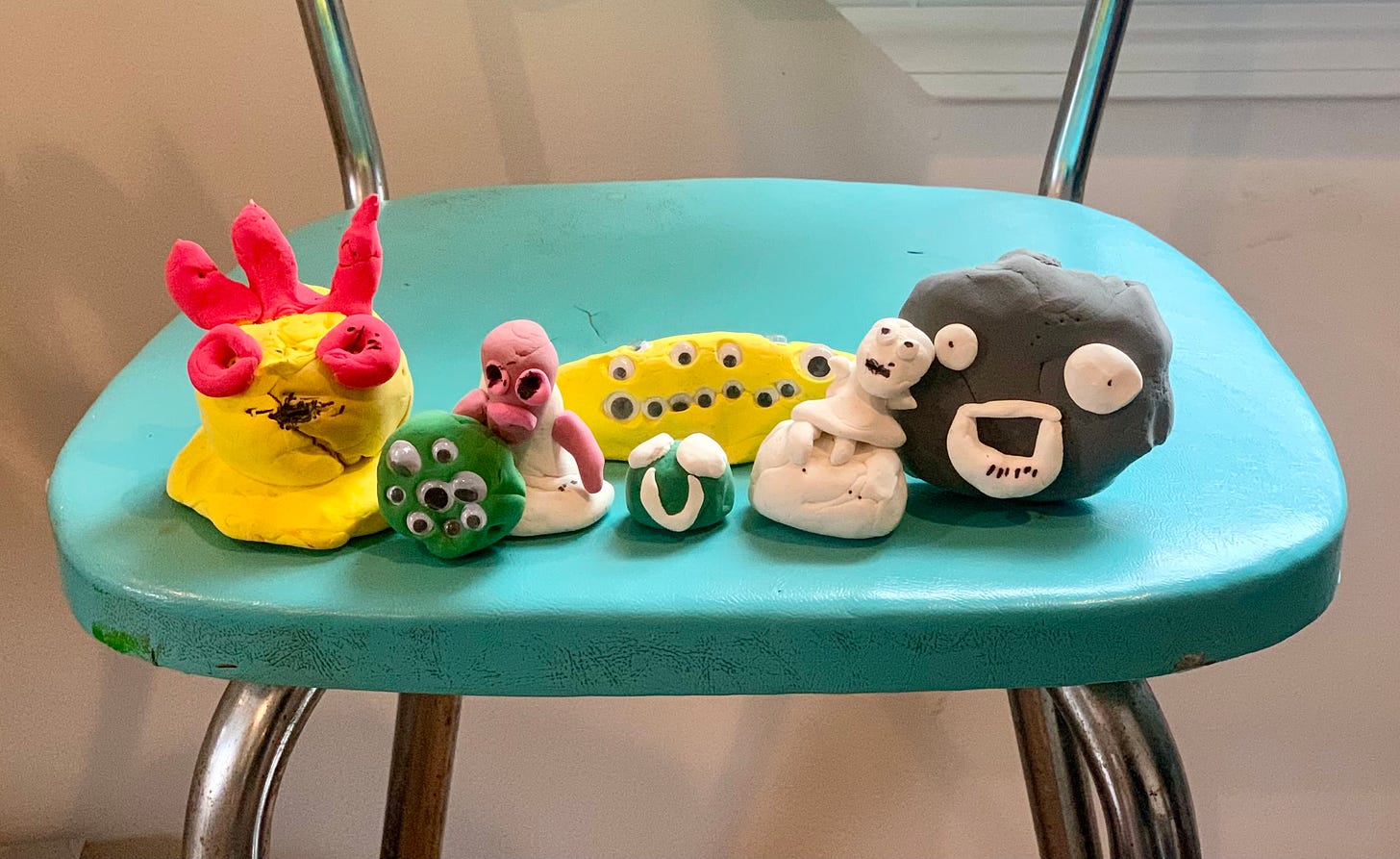 A line of Rebekah's sons clay creatures stand in a line on the teal blue chair Rebekah sat on as a child in her grandma's house. Each creature has it's own weird shape and expression. Some have of lots of eyes or googly eyes or two eyes of different sizes. Others have horns, some are frowning, others smiling, one seems to be sitting on a wheelchair. A line of Rebekah's sons clay creatures stand in a line on the teal blue chair Rebekah sat on as a child in her grandma's house. Each creature has it's own weird shape and expression. Some have of lots of eyes or googly eyes or two eyes of different sizes. Others have horns, some are frowning, others smiling, one seems to be sitting on a wheelchair.