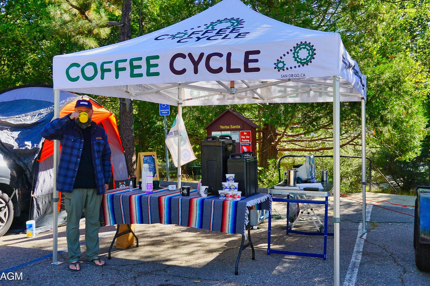 A bearded man drinks deeply from a yellow coffee mug while standing in front of a pop-up tent coffee shop in the parking lot of a campground. Pine trees fill the background and he's wearing a blue buffalo check coat, green hiking pants, and flip-flops.