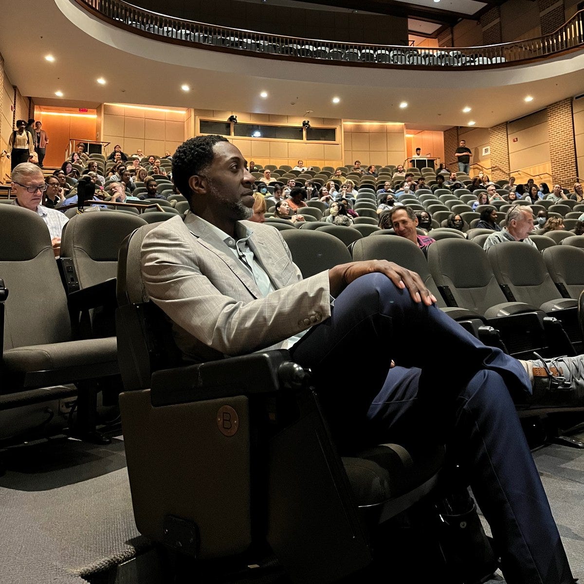 Nii Addy sitting in the front of an auditorium, wearing blue slacks and a grey blazer. Nii is sitting with his legs crossed, with a slight smile on his face.