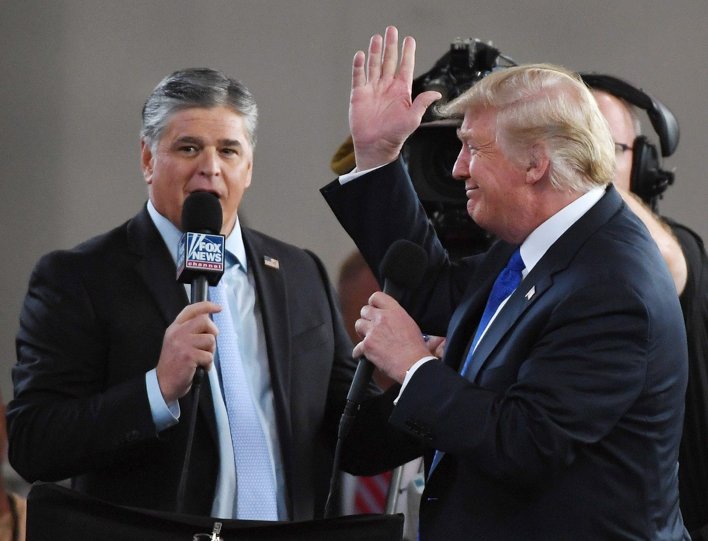 Two white men in suits stand on a stage, holding microphones. The one on the left has a mic branded "Fox News"