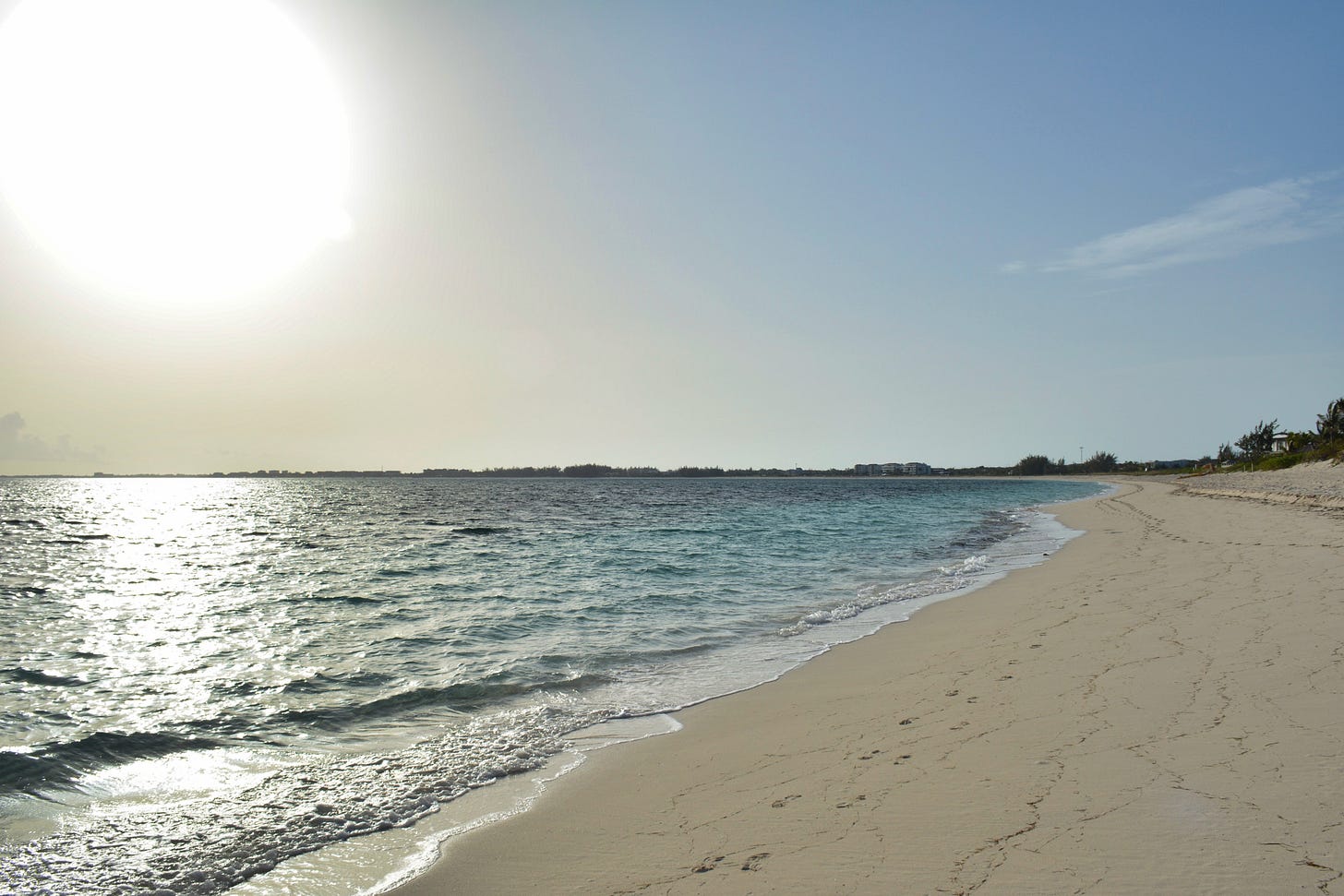 Sunrise on the berach with turquoise water, footprints in the sand, and the sun shining in the upper left corner