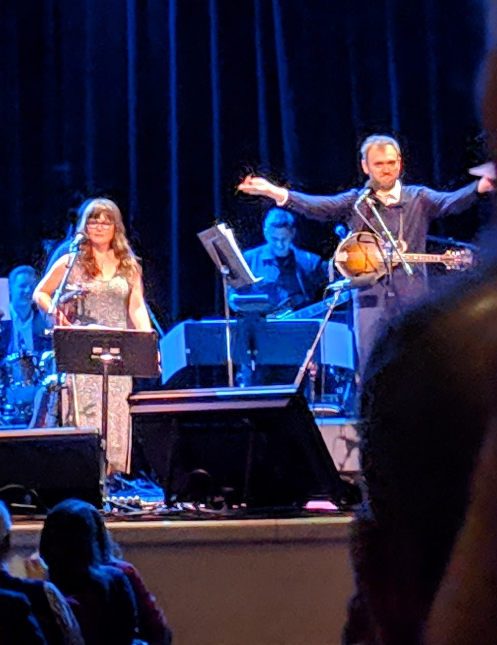 Sarah Watkins holding a violin next to Chris Thile gesturing with both arms wearing a mandolin