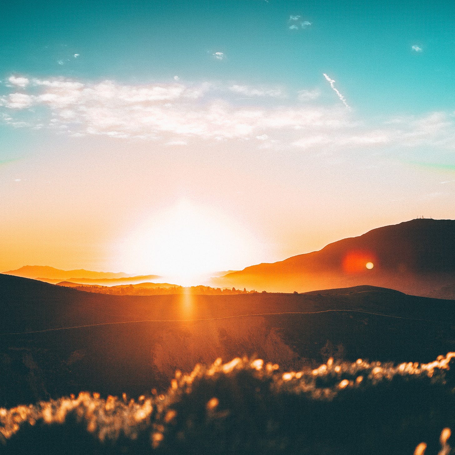 A brilliant aqua sky. Below are rolling hills. The bright orange sun is setting behind the hills, giving off a brilliant glow to everything around it.