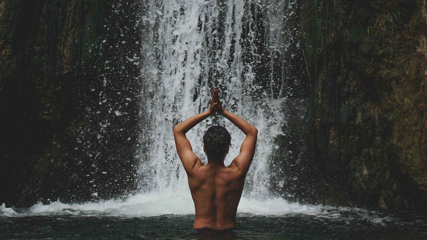 A naked person stands waist-deep in a pool at the bottom of a waterfall, faced toward the flow, palms pressed together in prayer-hands above the head.