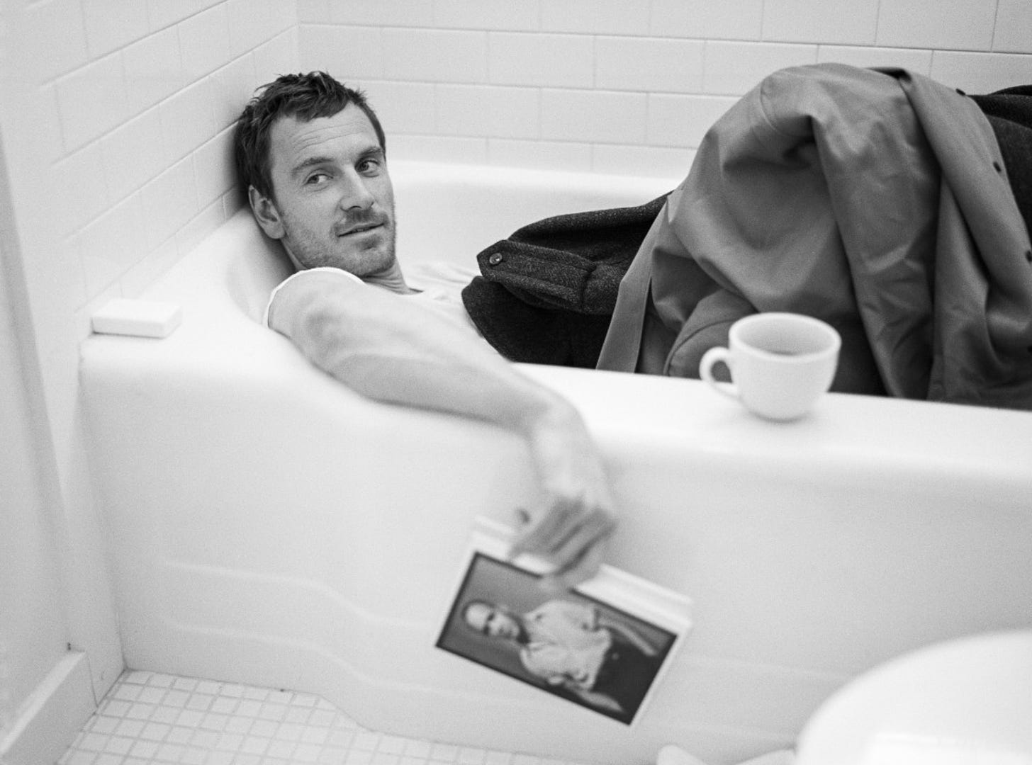 Michael Fassbender reclines clothed in a bathtub, holding a copy of a Genet book, for T Magazine