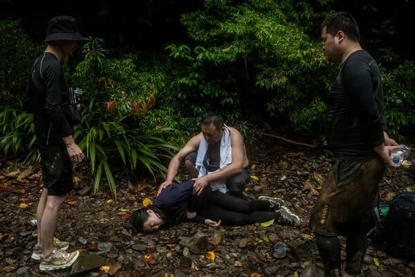 A young girl, lying on her side on the ground of a wooded area, clutches her stomach as a man with a shirt wrapped around his neck comforts her. Two other men are standing nearby.