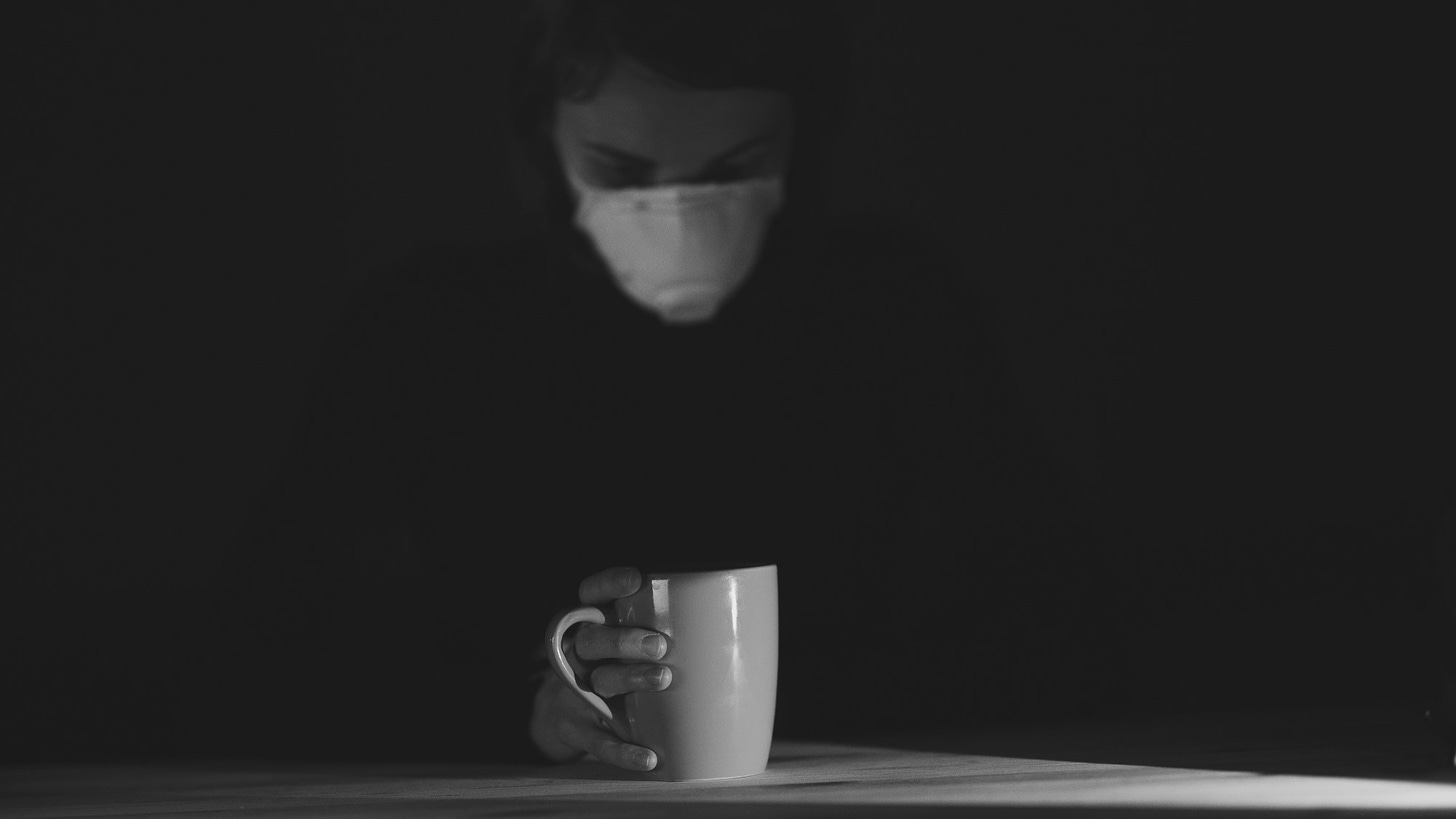 A person shrouded in darkness, wearing a mask, looks down at the cup of coffee they're holding