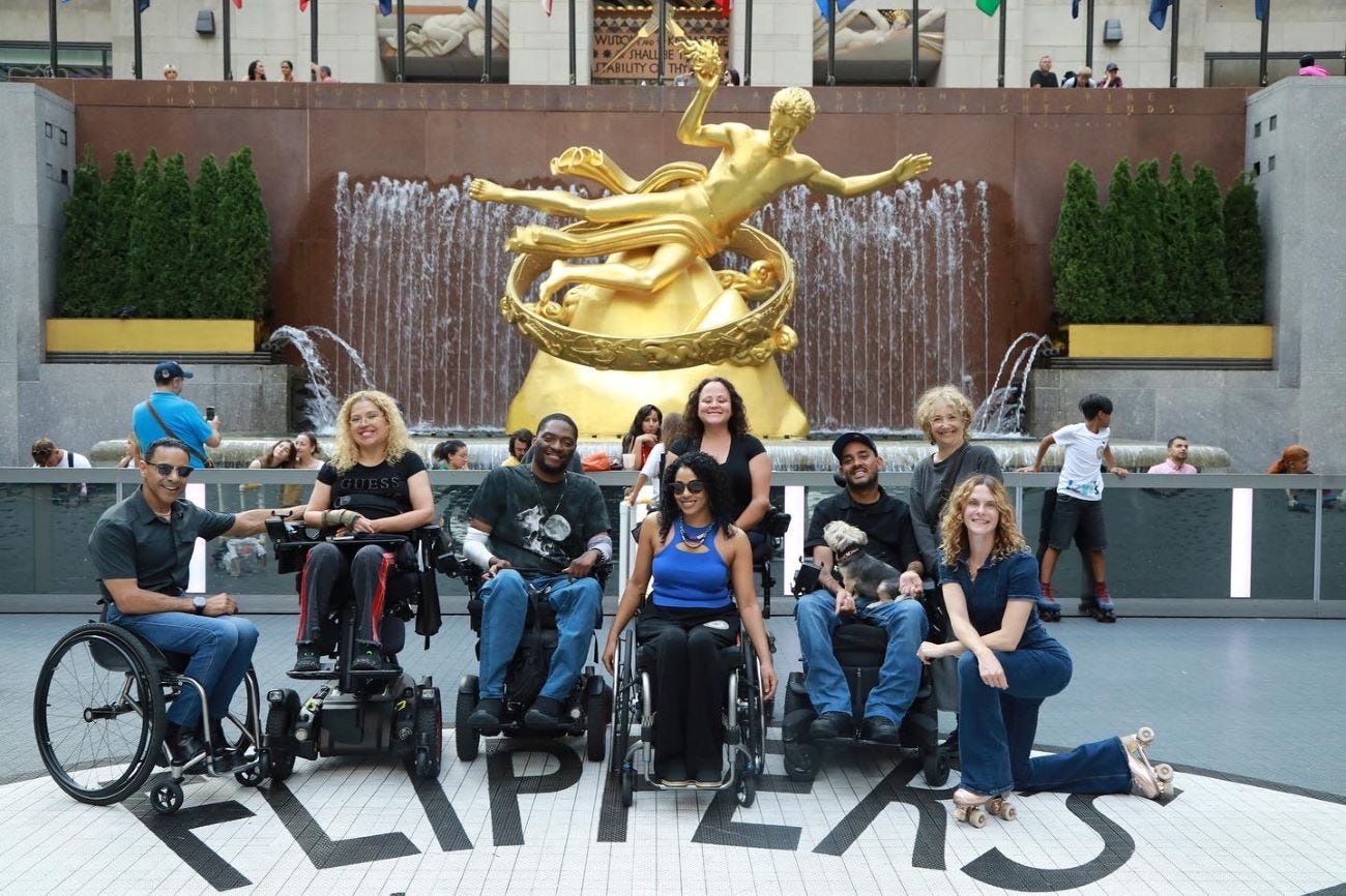 Organizers and artists, mostly wheelchair users, smile for a photo in front of the iconic gold sculpture in Rockefeller Center's roller/ice rink.