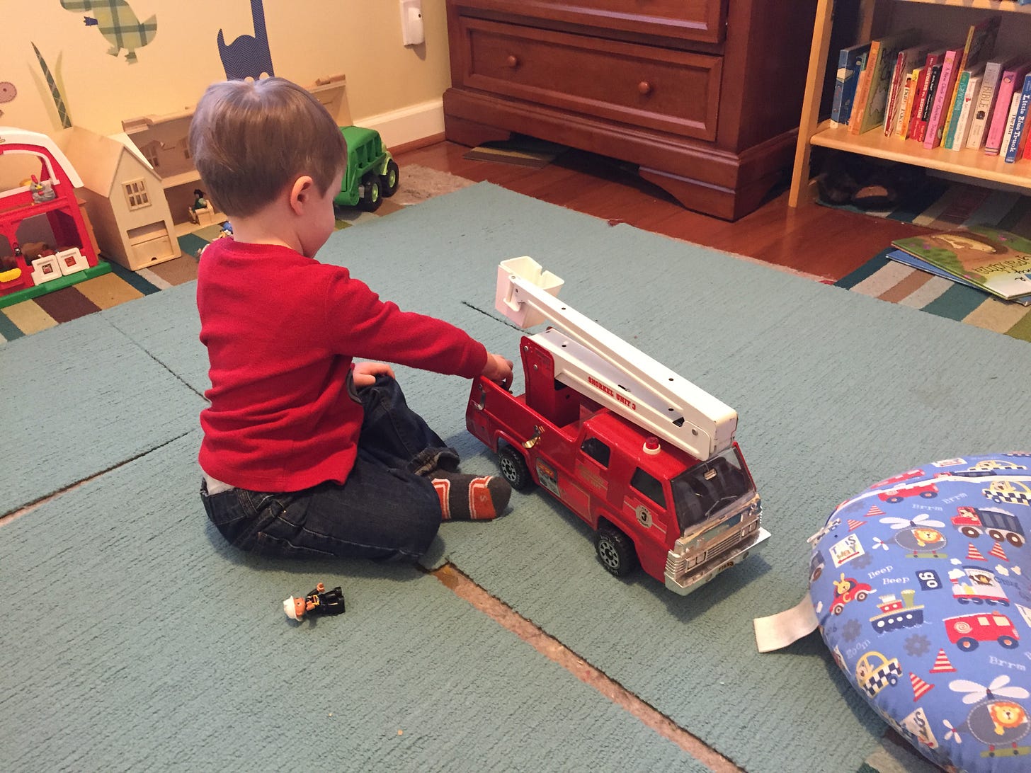 A white boy in a red sweater sitting on the floor and playing with a toy firetruck