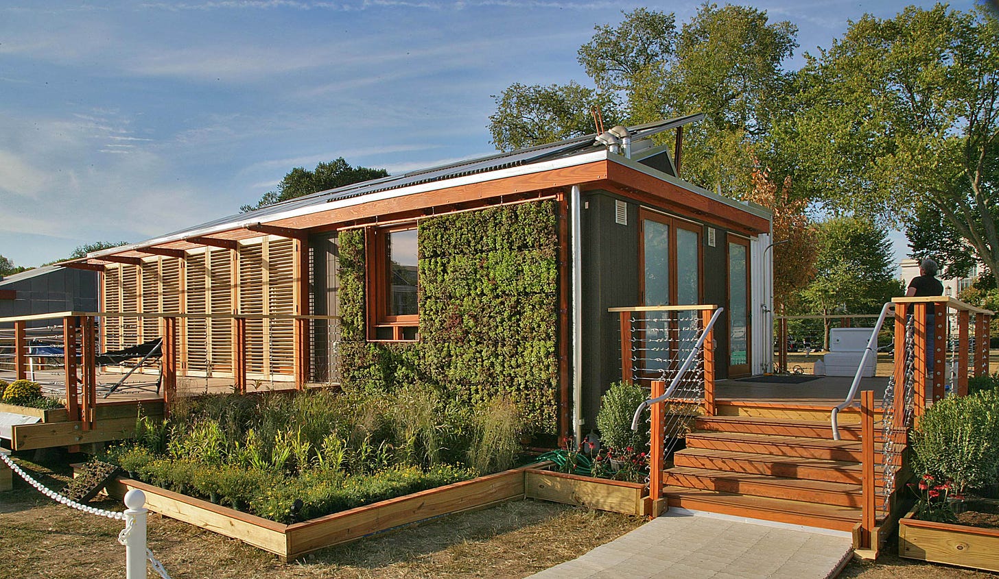 Photograph of a solar “tiny house,” LEAFHouse, the University of Maryland’s entry in the DOE’s 2007 Solar Decathlon competition. The view is from the corner of the house and you can see an outer wall covered with foliage, a section of wall with slatted shades, an open deck space and a small wild-looking garden plot out front.