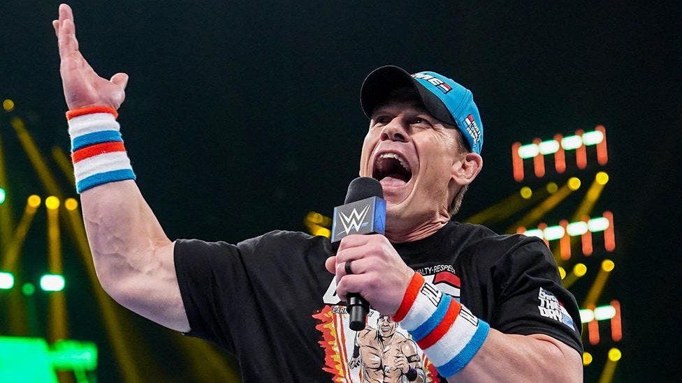 John Cena addressing the crowd in the wrestling ring. He is wearing a black t-shirt and holding a black microphone with the WWE logo on it in his left hand. He is wearing a light blue cap and has red, white and blue sweat bands on both wrists. His right arm is raised to hte sky.
