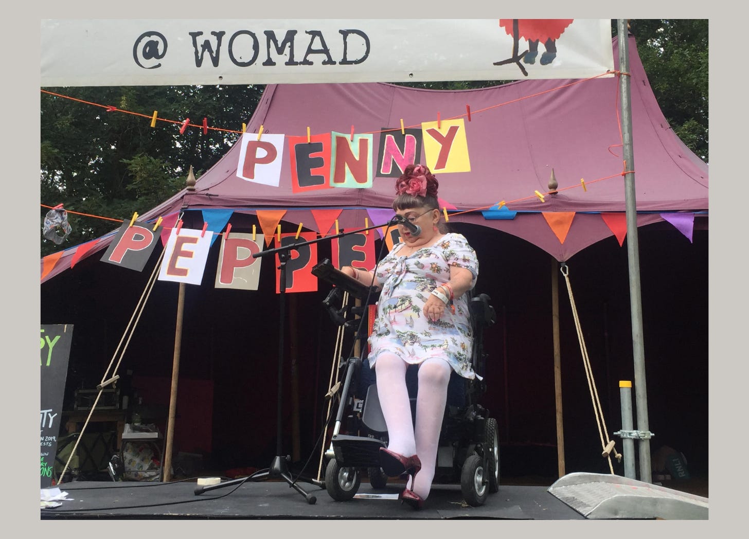 Penny is on a small outdoor stage in her wheelchair wearing a summery dress, white with pastel patterns. She has her red hair in a huge high bun, wears thigh high white socks and red stiletto heel shoes. Behind Penny, her name is spelled out with colourful letters through pegs on a line, and behind that is a low shaped dusky pink tent, the back stage area. Penny is looking down, sitting in front of the microphone, ready to speak.